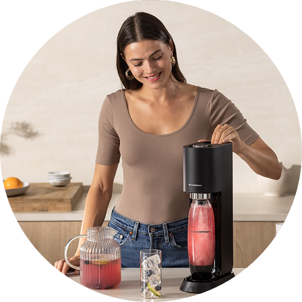 Kobieta w beżowej bluzce obsługująca czarną maszynę SodaStream z różowym napojem w butelce, obok dzbanek i szklanka z lodem