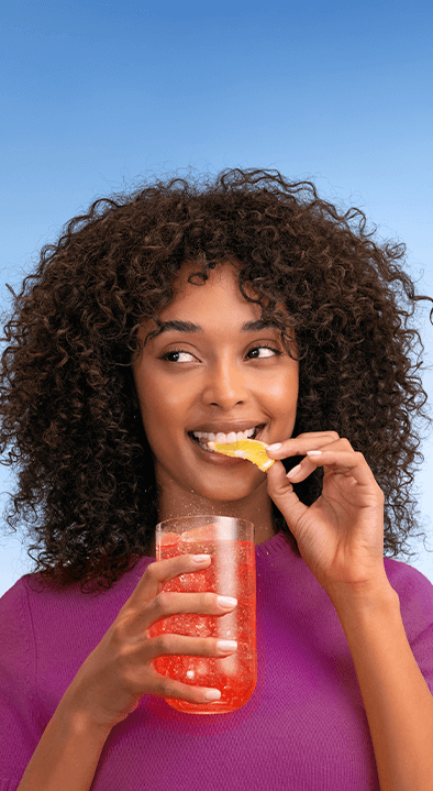 Dwie osoby wznoszą toast kieliszkami z gazowanymi napojami, jeden z plasterkiem brzoskwini, drugi z cytryną, na jasnym tle.